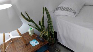 A neatly arranged hotel room with a wooden nightstand, a lamp, a remote control, a plant, and a bed with white bedding. - Cozy apartment in Interlagos (São Paulo)