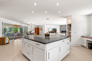 A modern kitchen with a large island, white cabinetry, and a view of the outdoors. - August House - Bold Design, Poolside Fun, Spa, Fire Pit & Views (Palm Springs)
