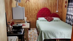 Pieni huone, jossa on sänky, pieni pöytä, jossa on vedenkeitin, tuuletin ja kuviollinen matto.