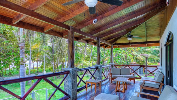 A covered outdoor area with wooden furniture, a view of greenery, and a security camera.