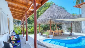 A covered outdoor area with a pool, wooden furniture, and a thatched-roof structure.