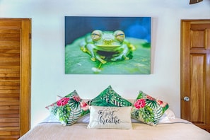 A bedroom with a bed, pillows, a wall-mounted painting of a frog, and wooden doors.