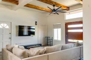 Una sala de estar con un sofá grande, un televisor de pantalla plana, un ventilador de techo y una ventana con persianas.