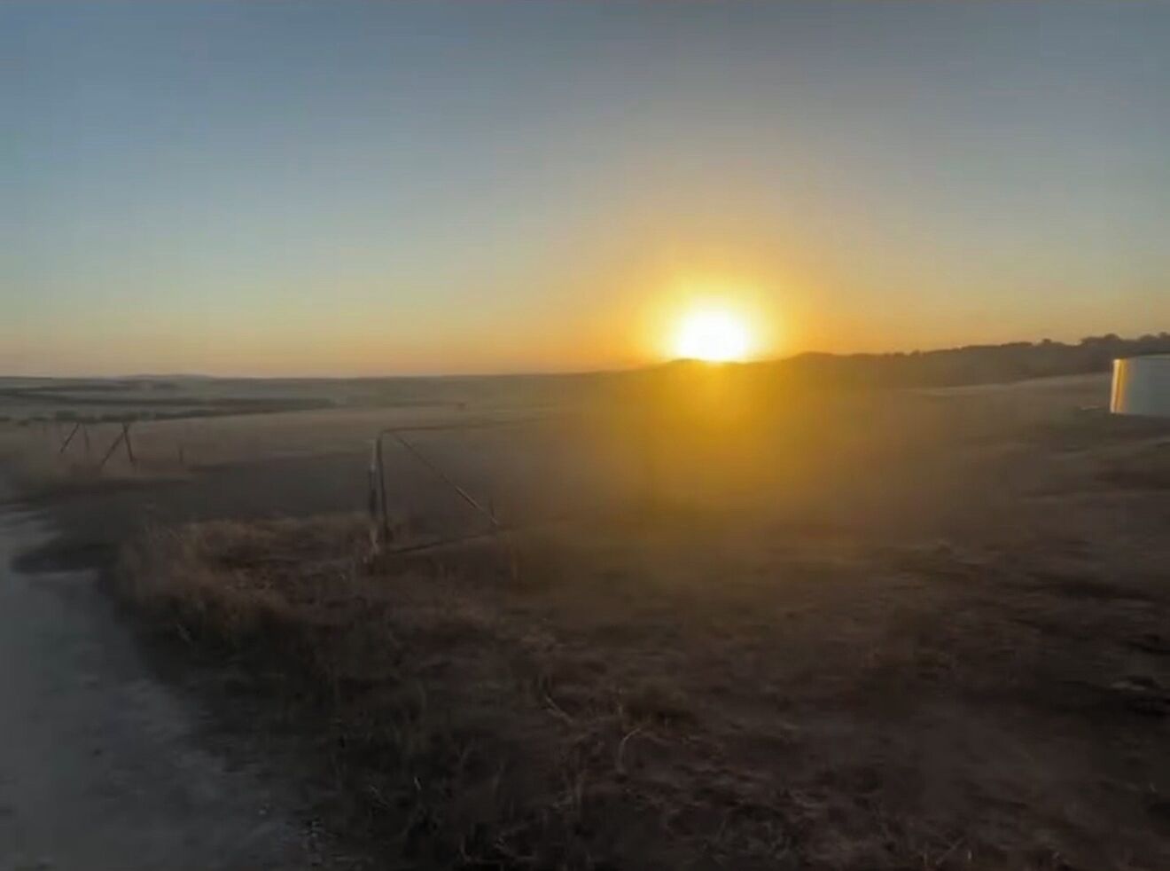 Een zonsondergang boven een vlak landschap met schaarse vegetatie en een heldere lucht.