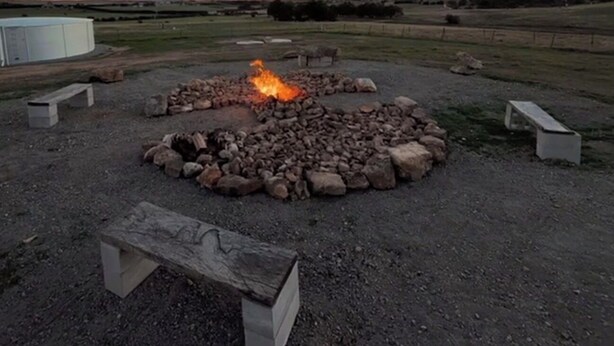 A circular stone fire pit with benches around it, set in an open field with a clear sky.
