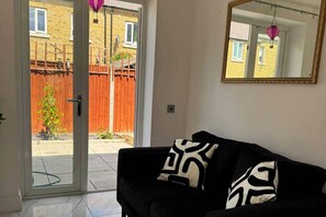 Ein Zimmer mit einer schwarzen Couch, zwei gemusterten Kissen, einem großen Spiegel und Blick durch eine Glastür auf einen Außenbereich mit Holzzaun und einer hängenden Laterne.