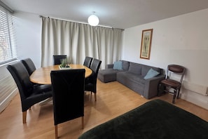 Ein Wohnzimmer mit Esstisch, Stühlen, einer Couch und einem grünen Hocker.
