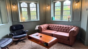 Une pièce comprenant un fauteuil en cuir, un canapé capitonné, une table basse en bois et deux fenêtres avec des lampes fixées au mur.