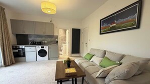 A living room with a sofa, a wooden coffee table, a kitchen area with appliances, and a painting on the wall. - 1 Bed - Sleeps 3 - Parking - Pets (London)