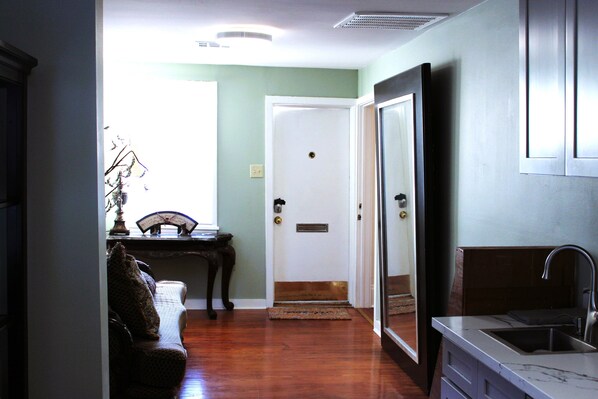 A hallway leading to a door with a doormat, a small table with a lamp, and a kitchen area with a sink and cabinets.