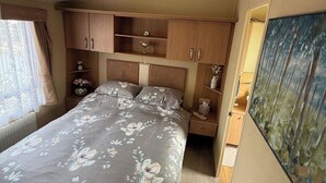 A bedroom with a bed, wooden cabinets, a nightstand, a vase, and a painting. - 2-Bed Caravan, Sleeps 6 w/ Parking, No Pass Needed (Abergele)