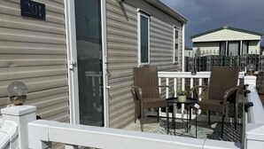 A balcony with a white railing, a sliding glass door, two wicker chairs, and a small table with a plant. - 2-Bed Caravan, Sleeps 6 w/ Parking, No Pass Needed (Abergele)