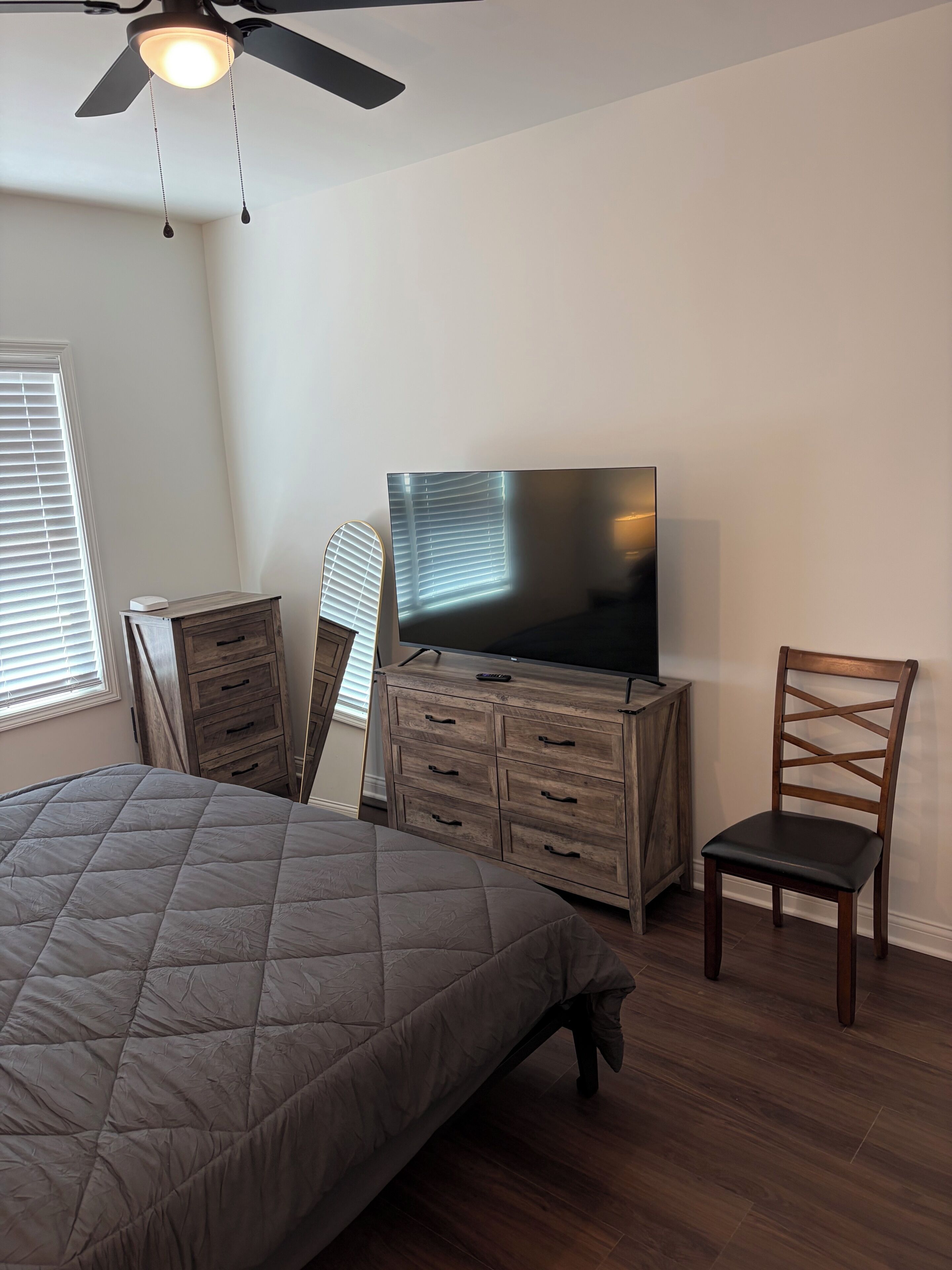Une chambre à coucher avec un lit, un téléviseur posé sur une commode en bois, un ventilateur de plafond et une fenêtre avec des stores.