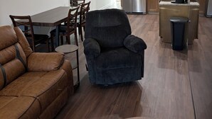 Una sala de estar moderna con un sofá, un sillón oscuro, una mesa de comedor con sillas, una zona de cocina con armarios de madera, un refrigerador de acero inoxidable y un cubo de basura.