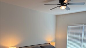 Un dormitorio con cama, mesitas de noche, ventilador de techo y ventana con persianas.
