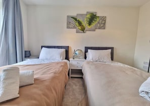 A hotel room with two beds, each with a white pillow and a beige blanket. There is a nightstand between the beds with a globe and a lamp. - Premium Central London Stay Near Sloane Square (London)