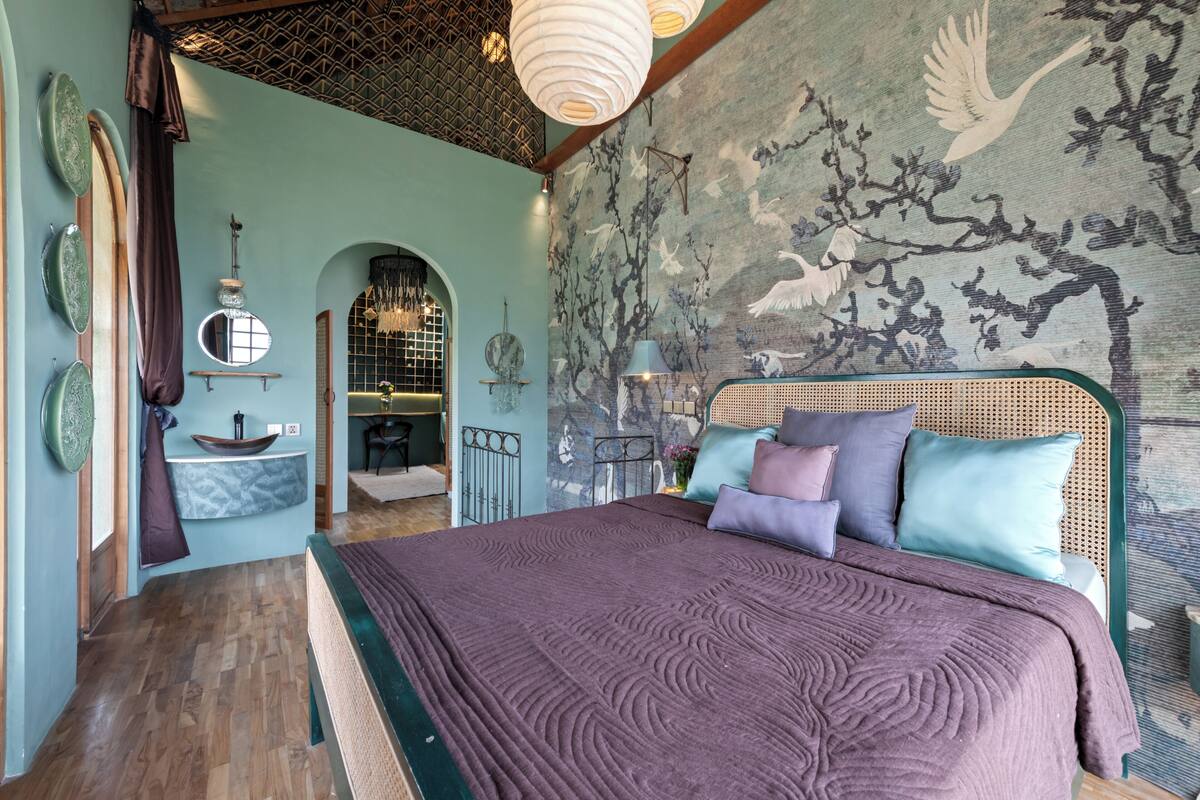 A bedroom with a bed, a mirror, a sink, and a wall with a tree and bird mural.