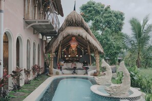 A pool with a decorative fountain, a bar area with seating, and a thatched-roof structure. - Atelier Ubud Art Lofts (Ubud)