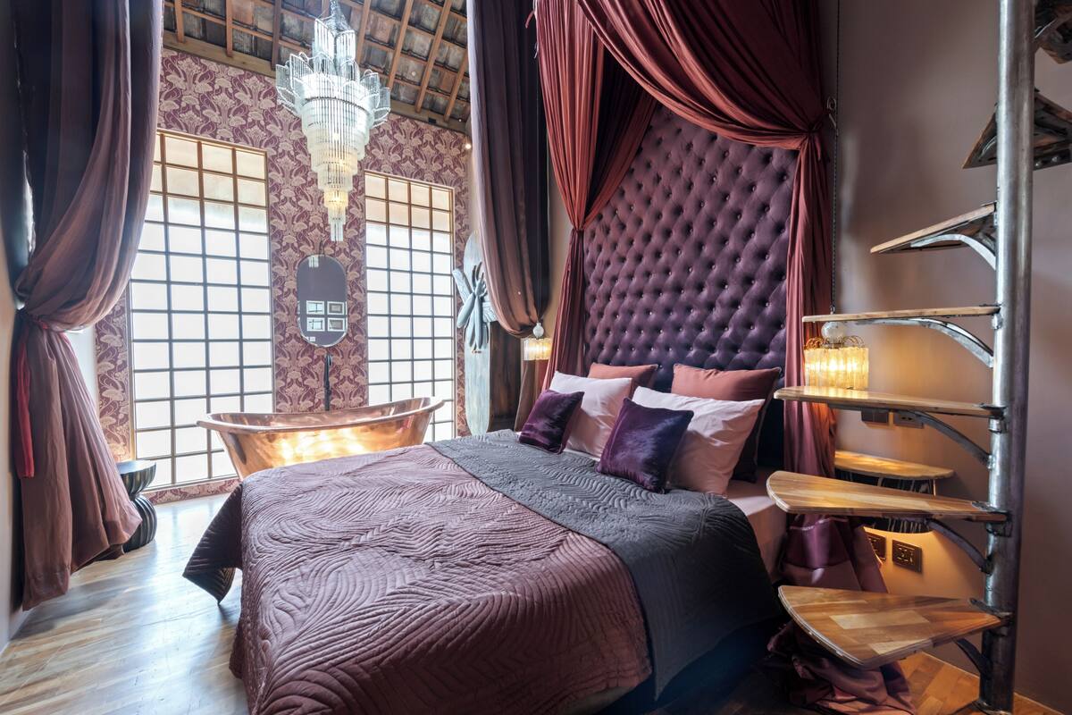 A bedroom with a large bed, a chaise lounge, a chandelier, and a unique staircase.