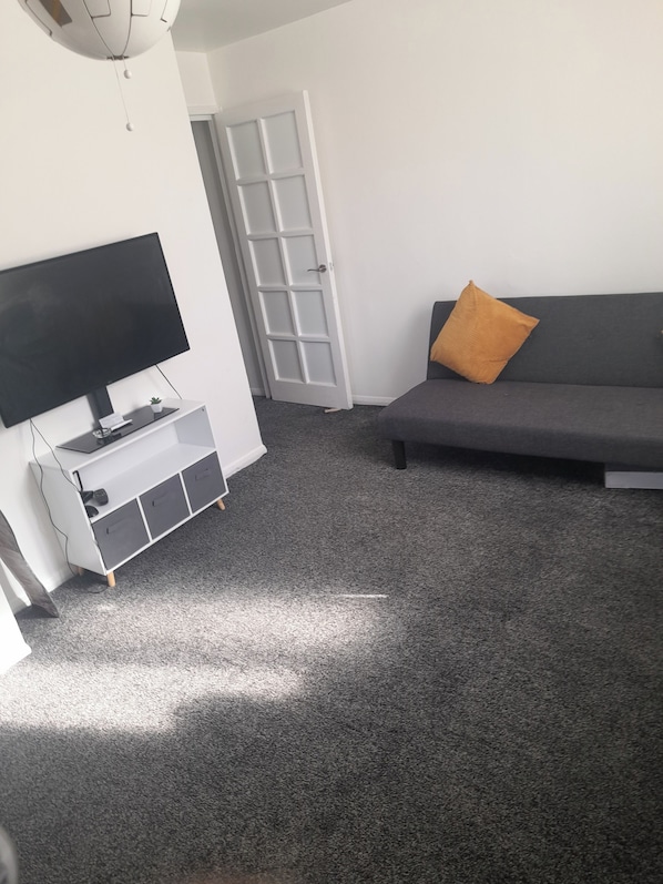 A living room with a flat-screen TV on a white stand, a gray sofa with an orange pillow, and a door leading to another room.
