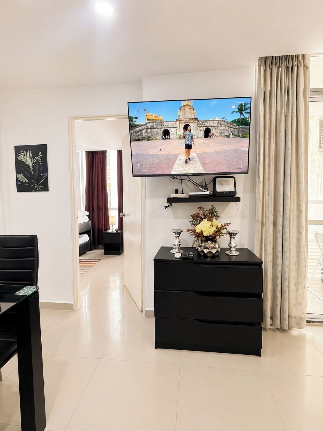 A living room with a flat-screen TV mounted on the wall, a black entertainment unit with decorative items, a dining area with a black table and chairs, and a view of a courtyard through a window.