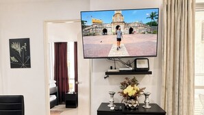 Een woonkamer met een flatscreen-tv aan de muur, een zwarte entertainmentkast met decoratieve objecten, een eethoek met een zwarte tafel en stoelen, en uitzicht op een binnenplaats door een raam.