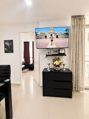 A living room with a flat-screen TV mounted on the wall, a black entertainment unit with decorative items, a dining area with a black table and chairs, and a view of a courtyard through a window. - Condominium in Cartagena (Cartagena de Indias)