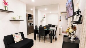 Een moderne woonkamer met een zwarte bank, een eetgedeelte met een tafel en stoelen, een aan de muur bevestigde televisie en een keuken op de achtergrond.