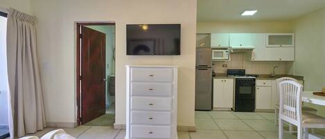 A hotel room with a bed, a white dresser, a television, a kitchen area with a microwave, stove, and cabinets.