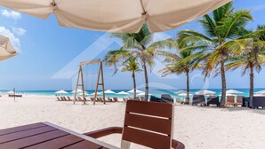 Une plage de sable blanc, des palmiers et une chaise en bois placée sous un parasol blanc.