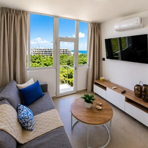 Ein modernes Wohnzimmer mit einer Couch, einem Couchtisch, einem Fernseher und einem Balkon mit Blick auf Grünflächen und Gebäude.