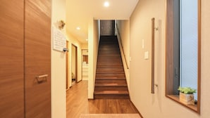 Un couloir avec un plancher en bois, un escalier, une porte en bois, un luminaire fixé au mur et une petite plante en pot.