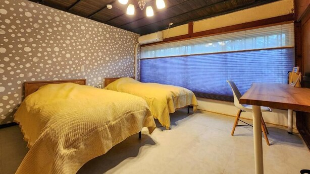 A room with two beds, a wooden ceiling, a window with blinds, and a small table. - taipi house  (Sado)