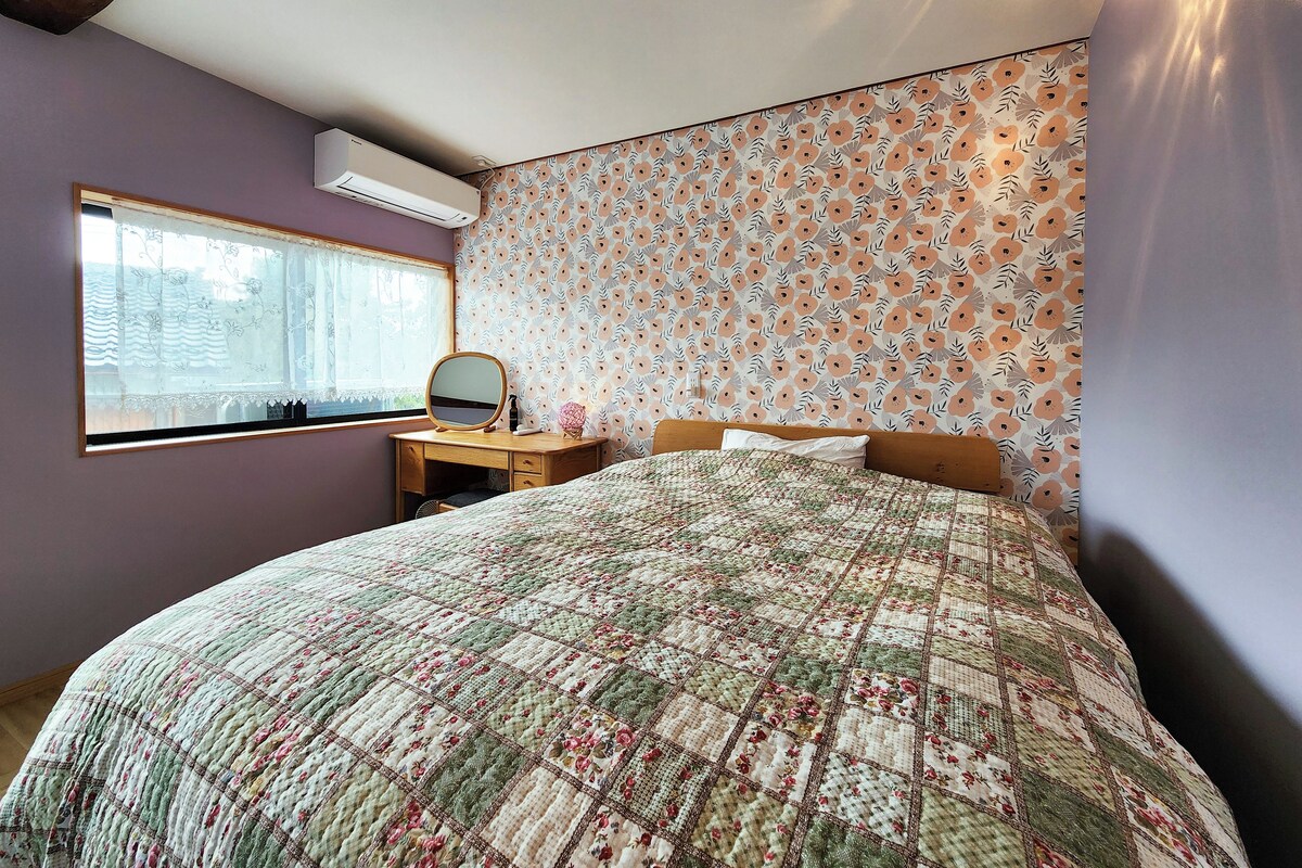A bedroom with a large bed, a wooden dresser, a mirror, and a window with a floral wallpaper pattern.