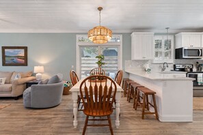 A modern kitchen with white cabinetry, a central island with bar stools, and a dining area with wooden chairs and a table. - Coastal Retreat on Beachview | Walk to Beach, Village + Family-Friendly Stay (Saint Simons Island)