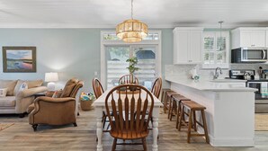 A modern kitchen with white cabinets, a central island, and a dining area with wooden chairs and a table. - Coastal Retreat on Beachview | Walk to Beach, Village + Family-Friendly Stay (Saint Simons Island)