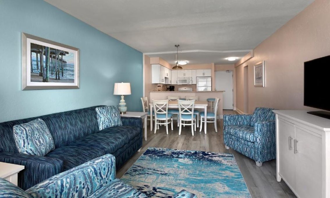A living room with a sofa, two armchairs, a coffee table, a TV, and a kitchen area with a dining table and chairs.