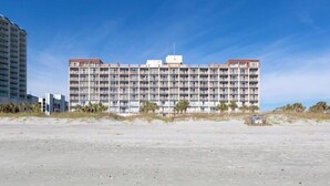 Ett hotell med flera våningar och en enhetlig fasad, beläget nära en strand med sparsam växtlighet.