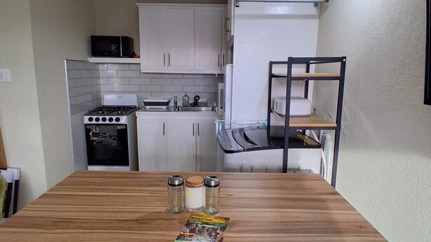 A compact kitchen with white cabinets, a microwave, a stove, and a small refrigerator. There is a wooden dining table with two chairs and a shelf with jars and a packet on it. - Cozy 1-bedroom apartment in fabulous Mullins, St. Peter with air conditioning! (Saint. Peter)