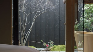 Une vue à travers une porte ouverte sur une pièce dont le sol est en bois, où l’on aperçoit une grande plante en pot, ainsi qu’un jardin avec une clôture en bois et un arbre.