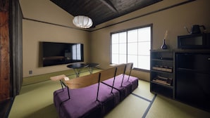 Une pièce avec un sol recouvert de tatamis, un plafond bas en bois, une télévision, une petite table, un banc et une fenêtre.