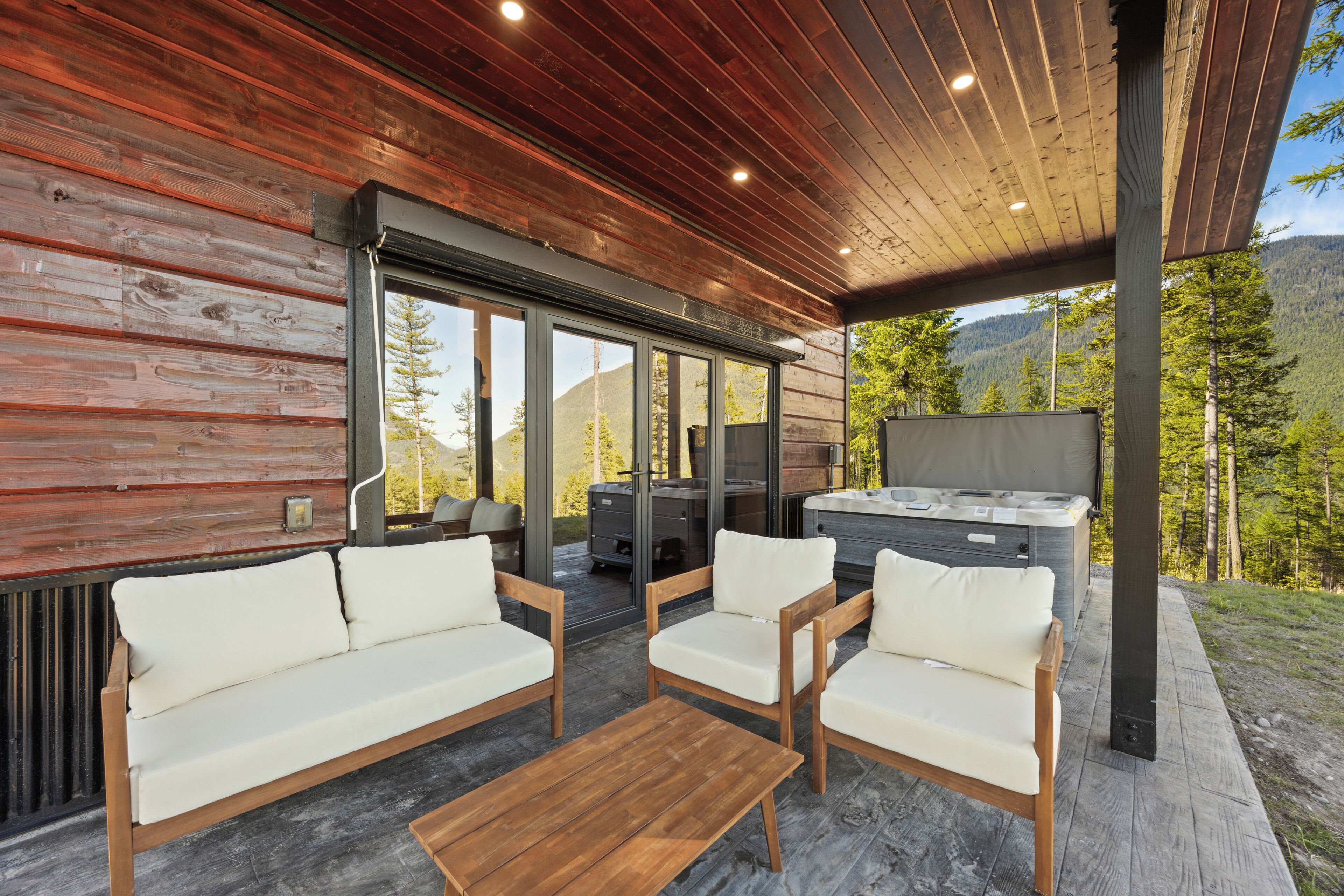 A covered outdoor seating area with wooden furniture, a hot tub, and a view of a mountainous landscape.