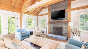 A spacious living room with a fireplace, wooden beams, and large windows. - Summerset Villa by Rove - Family Home With Pool & Hot Tub (Bridgehampton)