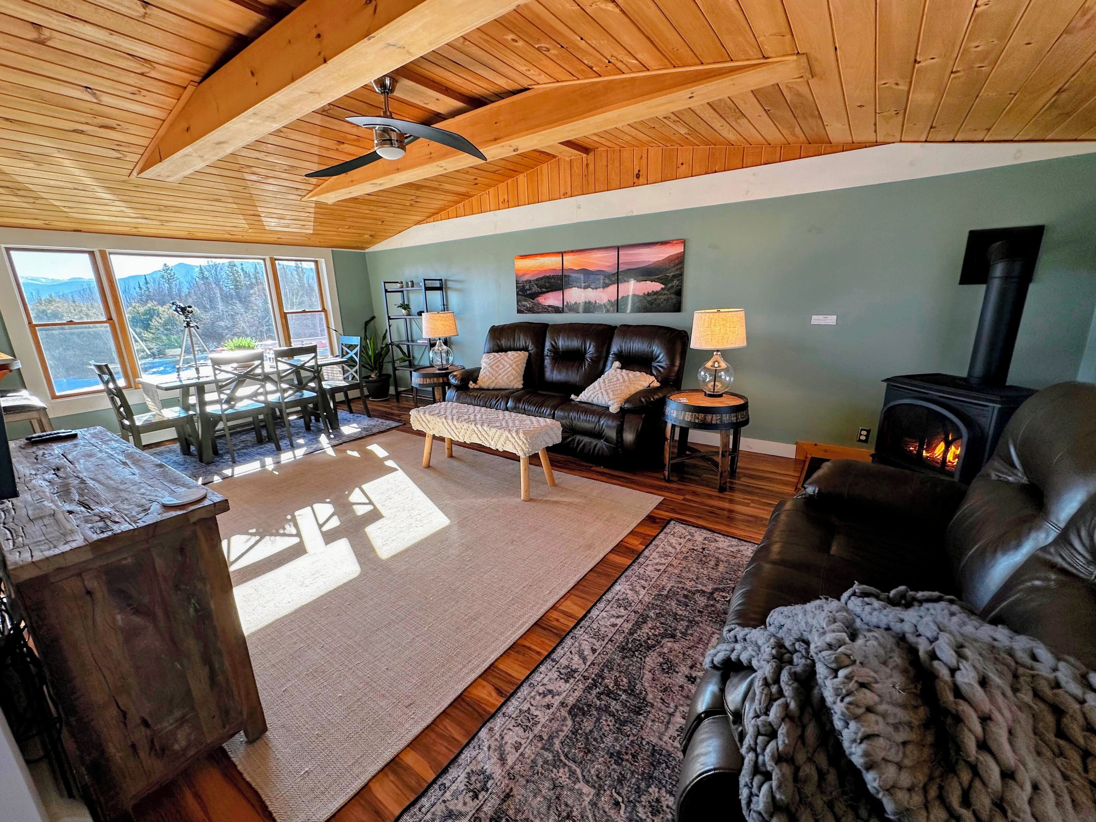 A cozy living room with a fireplace, leather sofas, a dining area with a table and chairs, and a large window offering a scenic view.