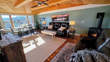 A cozy living room with a fireplace, leather sofas, a dining area with a table and chairs, and a large window offering a scenic view.