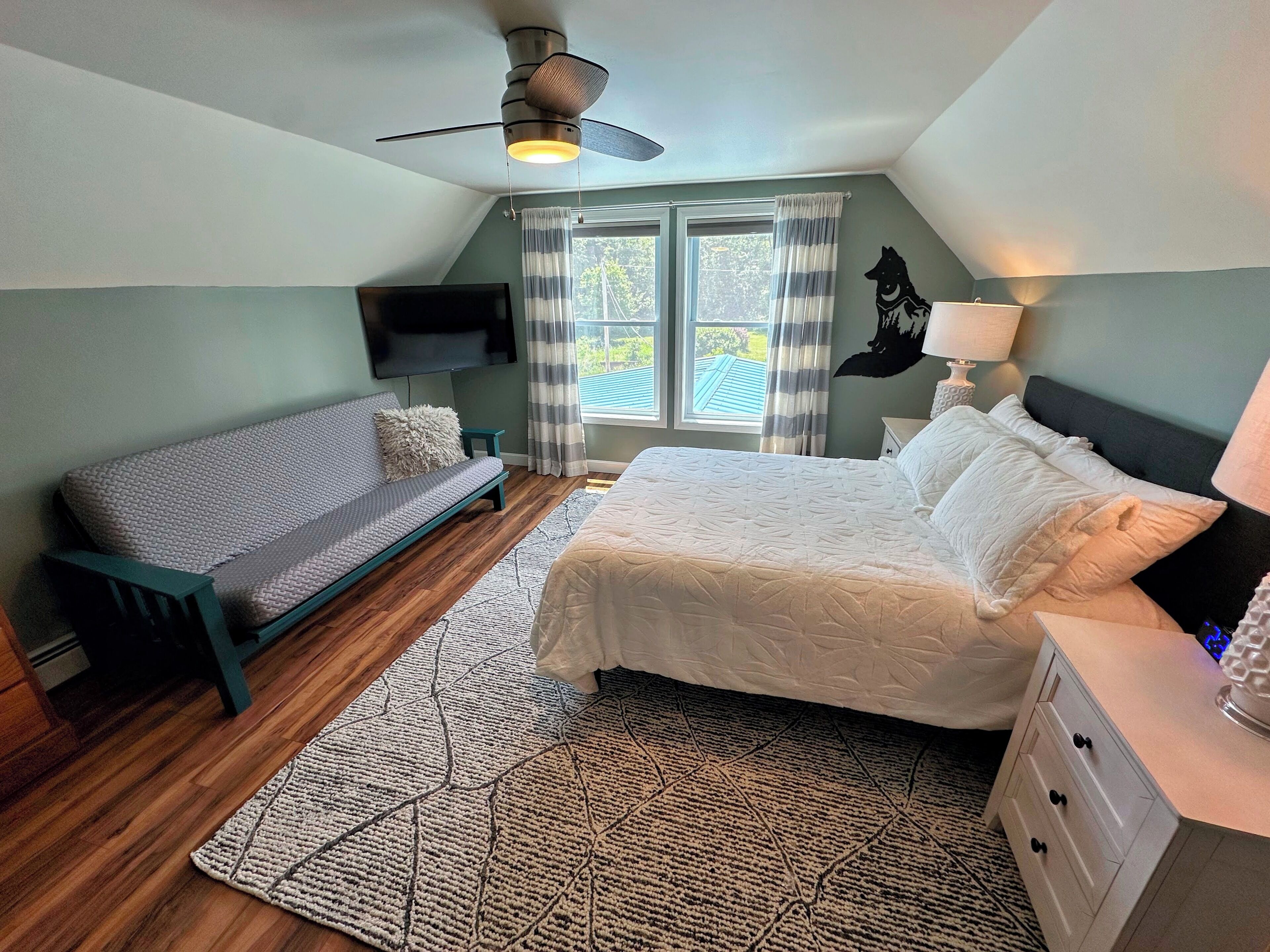 Une chambre à coucher comprenant un lit, un canapé, une télévision, un ventilateur de plafond et une fenêtre avec des rideaux.