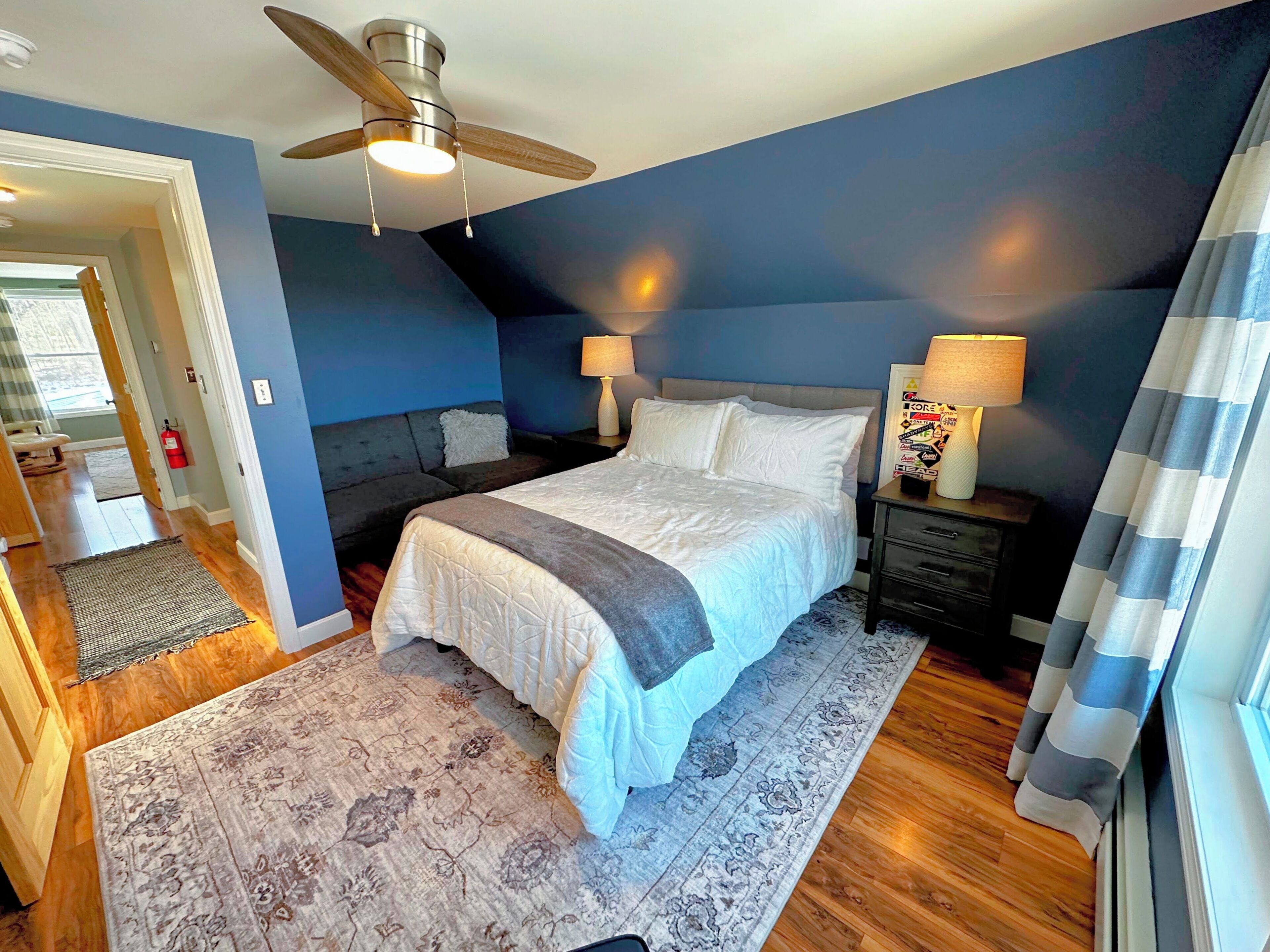 A bedroom with a bed, a ceiling fan, a nightstand with a lamp, and a window with curtains.