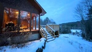 Eine Holzblockhütte mit einer überdachten Veranda, einer Treppe, die zu einer Terrasse führt, und einer verschneiten Landschaft im Hintergrund.
