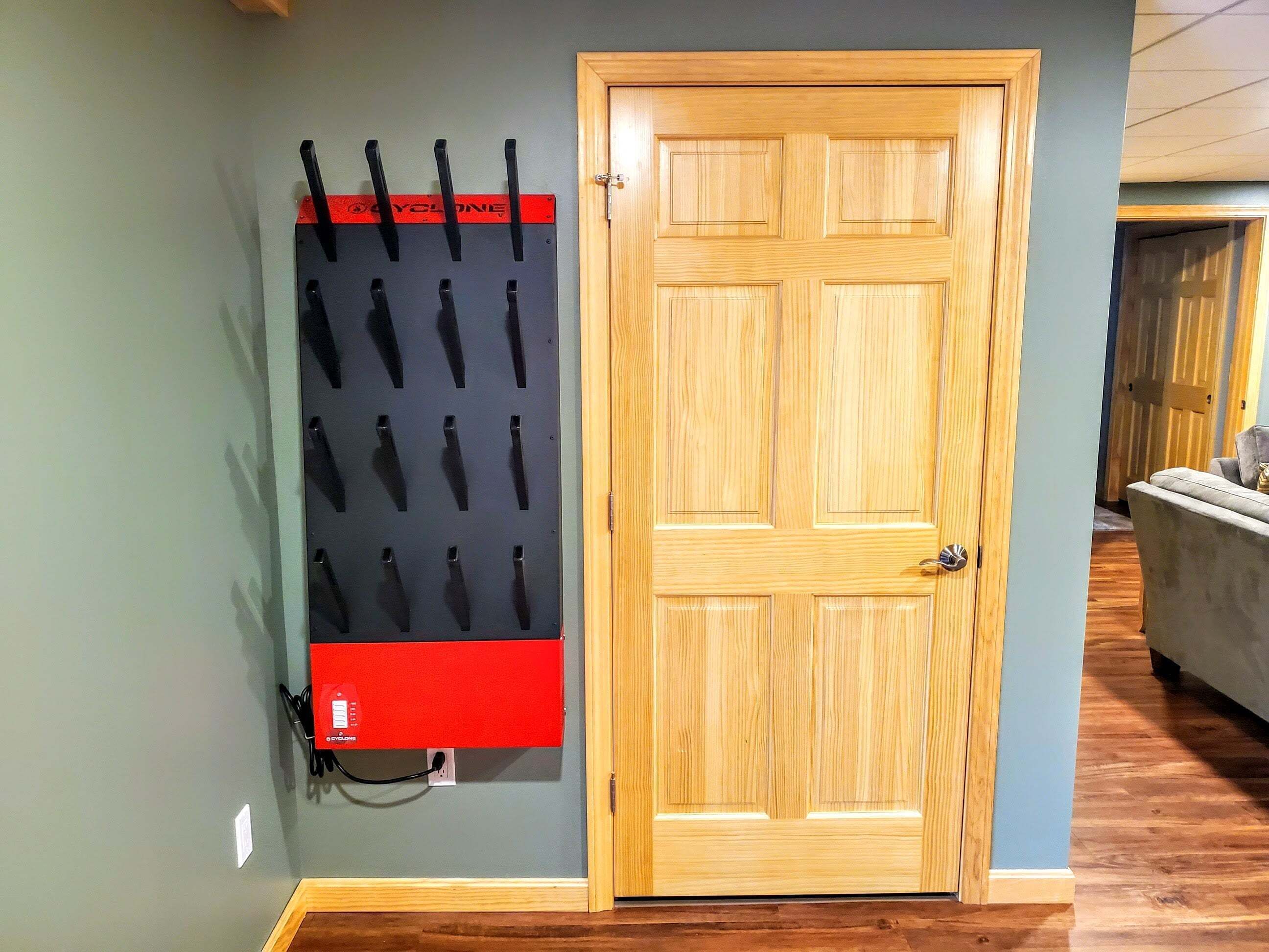 A wooden door with a black and red panel, a wall-mounted rack with black items, and a grey couch in the background.