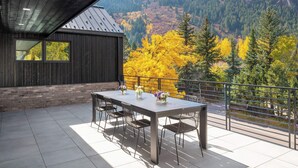 A modern outdoor dining area with a table and chairs, a wooden building with large windows, and a view of autumn-colored trees and mountains.
